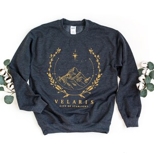 May include: A dark gray sweatshirt with a gold graphic design of a mountain range with a star and floral wreath. The text "Velaris City of Starlight" is printed below the graphic.