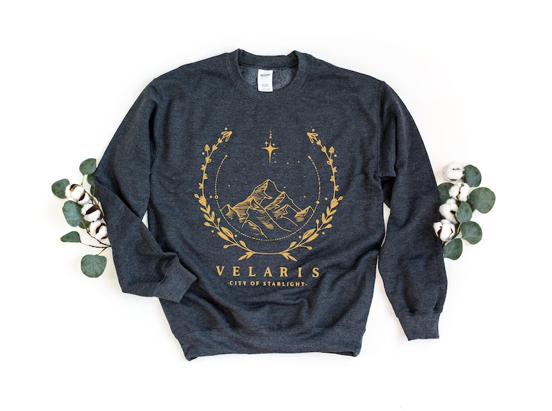 May include: A dark gray sweatshirt with a gold graphic design of a mountain range with a star and floral wreath. The text "Velaris City of Starlight" is printed below the graphic.