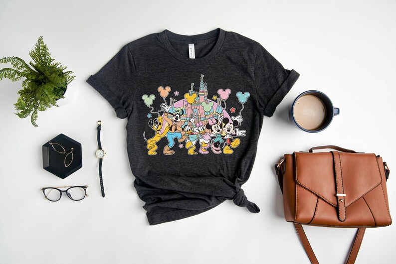 K&ouml;nnte beinhalten: Ein dunkelgraues T-Shirt mit einem bunten Motiv von Mickey Mouse, Minnie Mouse, Goofy und Pluto vor dem Schloss von Aschenputtel. Die Figuren halten Ballons und Sterne.