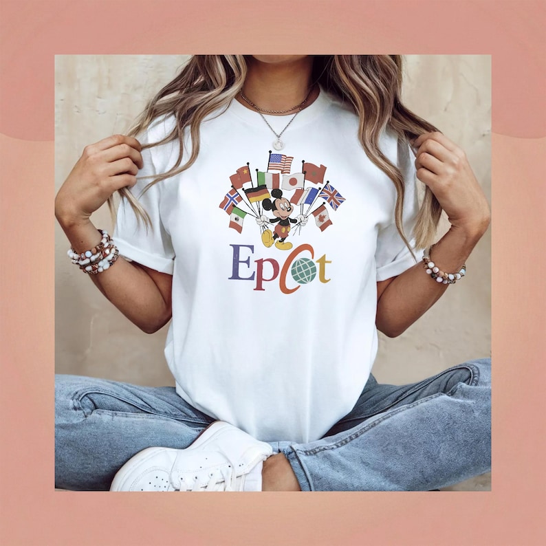 Op de afbeelding: Wit T-shirt met Mickey Mouse die vlaggen van verschillende landen vasthoudt. Het woord "Epcot" staat in kleurrijke letters eronder. Het T-shirt wordt gedragen met een blauwe spijkerbroek en witte sneakers.