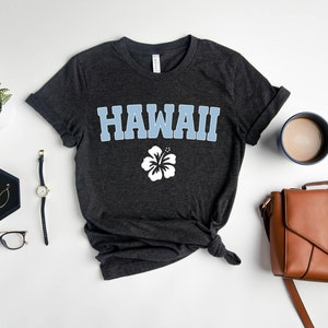 May include: A dark grey t-shirt with the word "HAWAII" printed in light blue letters and a white hibiscus flower underneath. The shirt is laid flat on a white surface with a watch, glasses, earrings, a coffee mug, and a brown leather purse.
