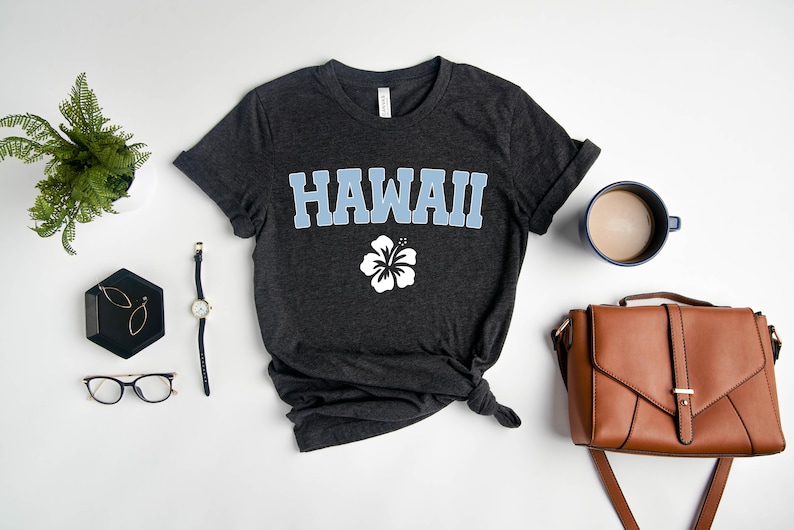 May include: A dark grey t-shirt with the word "HAWAII" printed in light blue letters and a white hibiscus flower underneath. The shirt is laid flat on a white surface with a watch, glasses, earrings, a coffee mug, and a brown leather purse.