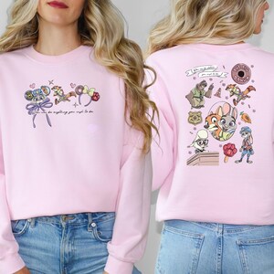 May include: A pale pink sweatshirt with cartoon illustrations and the text "You can be anything you wish to be." The front has character designs and a bow. The back has more character designs and a donut.