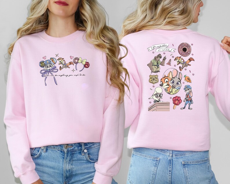 May include: A pale pink sweatshirt with cartoon illustrations and the text "You can be anything you wish to be." The front has character designs and a bow. The back has more character designs and a donut.