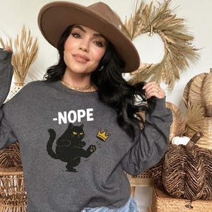 May include: Dark gray sweatshirt with the word "-NOPE" above a black cat kicking a gold crown. The cat has yellow eyes and is wearing a gold crown. The sweatshirt is a crew neck style.