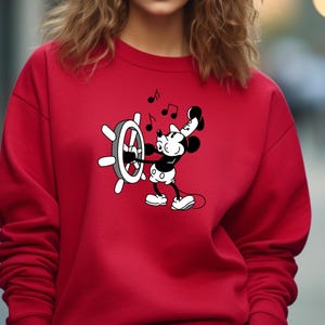 May include: Red sweatshirt featuring a black and white cartoon character, holding a ship's wheel and musical notes. The character is in a classic animated style. The sweatshirt is a solid red color.