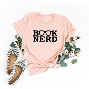 May include: A light pink t-shirt with the words "BOOK NERD" in black, with glasses above the word "NERD". The shirt is styled with a pair of striped flats and greenery. The shirt is made of a soft material.
