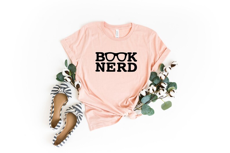 May include: A light pink t-shirt with the words "BOOK NERD" in black, with glasses above the word "NERD". The shirt is styled with a pair of striped flats and greenery. The shirt is made of a soft material.