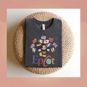 Op de afbeelding: Donkergrijs t-shirt met Mickey Mouse die vlaggen van verschillende landen vasthoudt. Het woord "Epcot" staat in kleurrijke letters eronder. Het shirt is opgevouwen op een geweven, ronde mat.