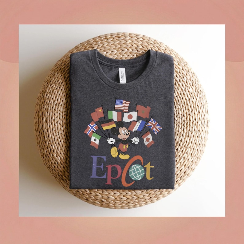 Op de afbeelding: Donkergrijs t-shirt met Mickey Mouse die vlaggen van verschillende landen vasthoudt. Het woord "Epcot" staat in kleurrijke letters eronder. Het shirt is opgevouwen op een geweven, ronde mat.