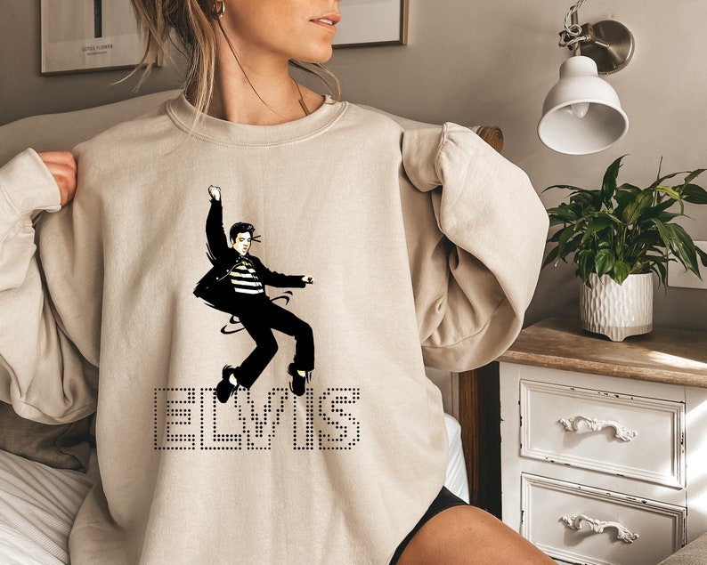 K&ouml;nnte beinhalten: Ein beiger Pullover mit einem Schwarzwei&szlig;bild von Elvis Presley. Das Bild zeigt Elvis in der Luft, mit ausgestreckten Armen und gekreuzten Beinen. Das Wort "ELVIS" ist in Schwarz unter dem Bild gedruckt.