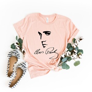 Pode incluir: Camiseta rosa claro com uma ilustra&ccedil;&atilde;o preta do rosto e assinatura de Elvis Presley. A imagem &eacute; estilizada com sapatilhas de la&ccedil;o listradas e galhos de algod&atilde;o e vegeta&ccedil;&atilde;o, criando uma est&eacute;tica casual e art&iacute;stica.