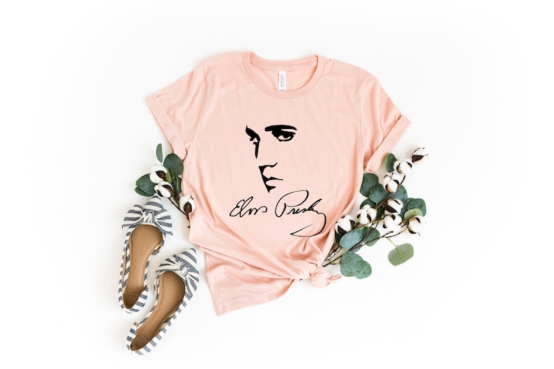 Pode incluir: Camiseta rosa claro com uma ilustra&ccedil;&atilde;o preta do rosto e assinatura de Elvis Presley. A imagem &eacute; estilizada com sapatilhas de la&ccedil;o listradas e galhos de algod&atilde;o e vegeta&ccedil;&atilde;o, criando uma est&eacute;tica casual e art&iacute;stica.