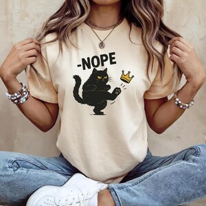 May include: A light beige t-shirt featuring a black cat kicking a gold crown. The word "-NOPE" is printed above the cat. The cat has yellow eyes and is kicking the crown with its paw. The shirt is a casual style.
