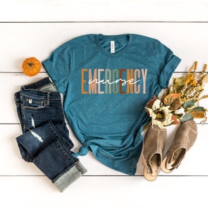Puede incluir: Una camiseta de manga corta color verde azulado con la palabra EMERGENCY en letras de bloque en varios tonos tierra y la palabra nurse en cursiva blanca en el centro. La camiseta se combina con unos vaqueros azul oscuro desgastados.