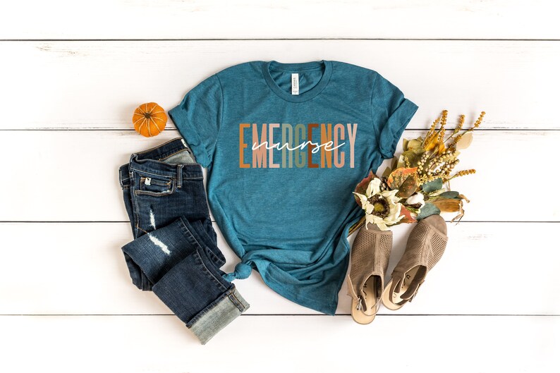 Puede incluir: Una camiseta de manga corta color verde azulado con la palabra EMERGENCY en letras de bloque en varios tonos tierra y la palabra nurse en cursiva blanca en el centro. La camiseta se combina con unos vaqueros azul oscuro desgastados.