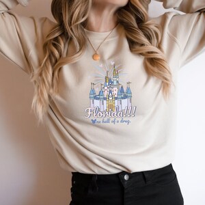 May include: Beige sweatshirt featuring a colorful illustration of a castle with the text "Florida!!!" and "...a hell of a drug." The design includes blue, pink, and yellow accents. The sweatshirt is a casual style.