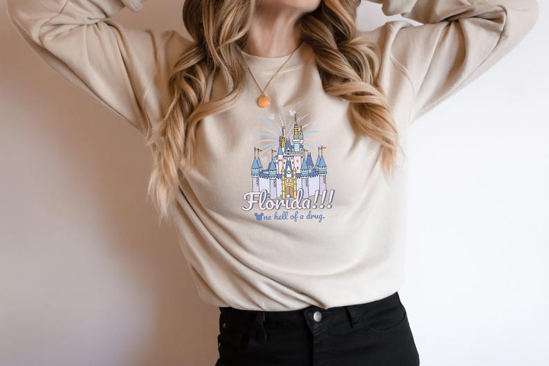 May include: Beige sweatshirt featuring a colorful illustration of a castle with the text "Florida!!!" and "...a hell of a drug." The design includes blue, pink, and yellow accents. The sweatshirt is a casual style.