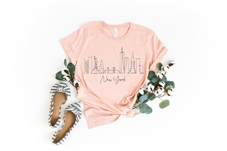 Peut inclure: Un t-shirt rose avec un dessin au trait noir de l'horizon de New York et le texte "New York". Le t-shirt est pos&eacute; &agrave; plat sur un fond blanc avec une branche d'eucalyptus et des boules de coton.