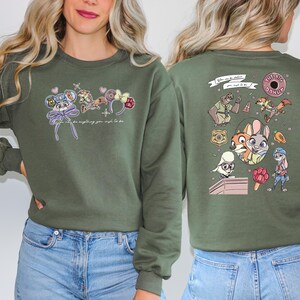 May include: An olive green sweatshirt with cartoon character designs on the front and back. The front has character-themed accessories and the text "Can be anything you wish to be." The back features character illustrations and the text "The Big Donut."