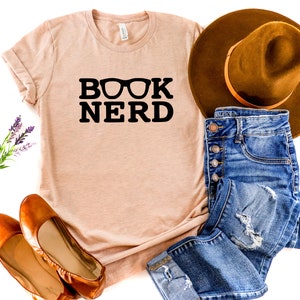 May include: A light peach-colored t-shirt with the words "BOOK NERD" in bold black letters, with eyeglasses replacing the "O". The shirt is paired with blue jeans, a brown hat, and brown flats.