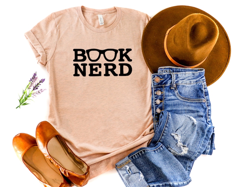 May include: A light peach-colored t-shirt with the words "BOOK NERD" in bold black letters, with eyeglasses replacing the "O". The shirt is paired with blue jeans, a brown hat, and brown flats.