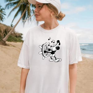 May include: White t-shirt featuring a black and white cartoon of Mickey Mouse at a ship's wheel with musical notes. The shirt is a casual style, suitable for everyday wear. The background shows a beach scene.