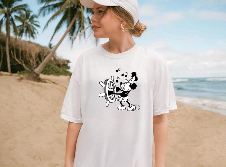 May include: White t-shirt featuring a black and white cartoon of Mickey Mouse at a ship's wheel with musical notes. The shirt is a casual style, suitable for everyday wear. The background shows a beach scene.