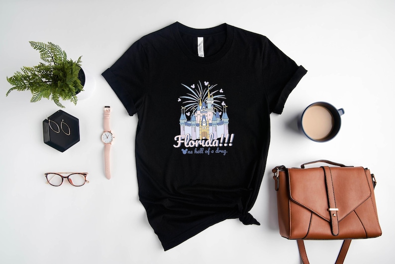 May include: Black t-shirt with a graphic of a fairytale castle, fireworks, and the text "Florida!!! no hell of a drug." Accessories include a watch, earrings, glasses, a plant, a coffee cup, and a brown purse.