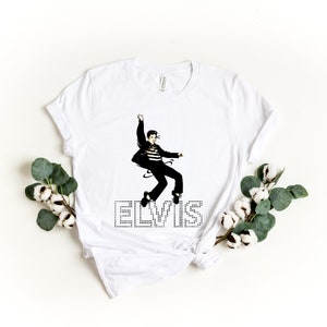 K&ouml;nnte beinhalten: Wei&szlig;es T-Shirt mit einem Schwarzwei&szlig;bild von Elvis Presley in einer Sprungpose. Das Wort "ELVIS" ist in Schwarz mit wei&szlig;en Punkten unter dem Bild gedruckt.