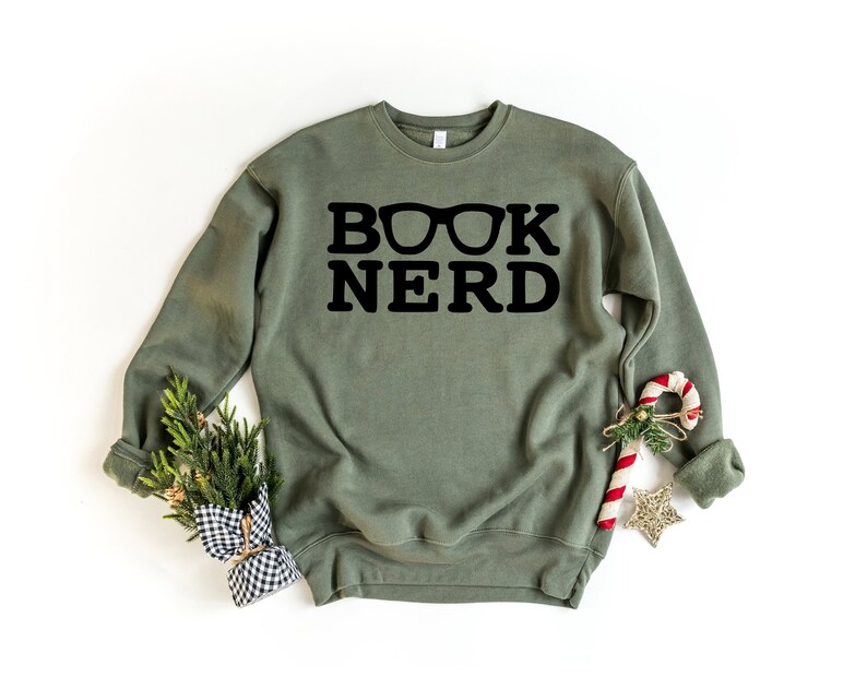 May include: An olive green sweatshirt with the words "BOOK NERD" in bold black letters, with glasses incorporated into the design. The sweatshirt is displayed on a white background, with holiday decorations including a candy cane and a small evergreen bundle.