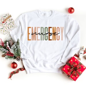 Puede incluir: Una sudadera blanca de manga larga con cuello redondo que presenta la palabra EMERGENCY en letras grandes y multicolores, con la palabra nurse escrita en una fuente cursiva negra superpuesta en el centro del texto.