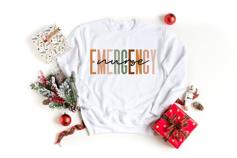 Puede incluir: Una sudadera blanca de manga larga con cuello redondo que presenta la palabra EMERGENCY en letras grandes y multicolores, con la palabra nurse escrita en una fuente cursiva negra superpuesta en el centro del texto.