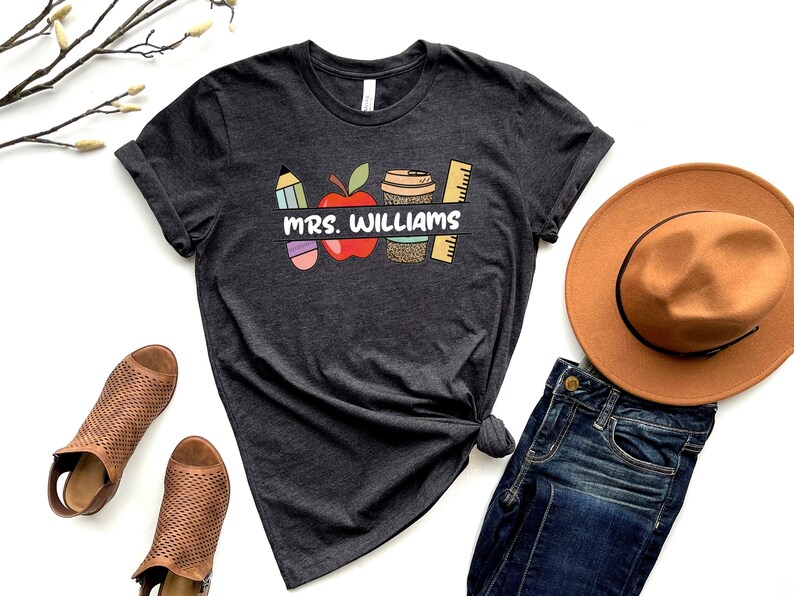 May include: A dark gray t-shirt with a graphic design of a red apple, a pencil, a coffee cup, and a ruler. The text "Mrs. Williams" is printed on the shirt.