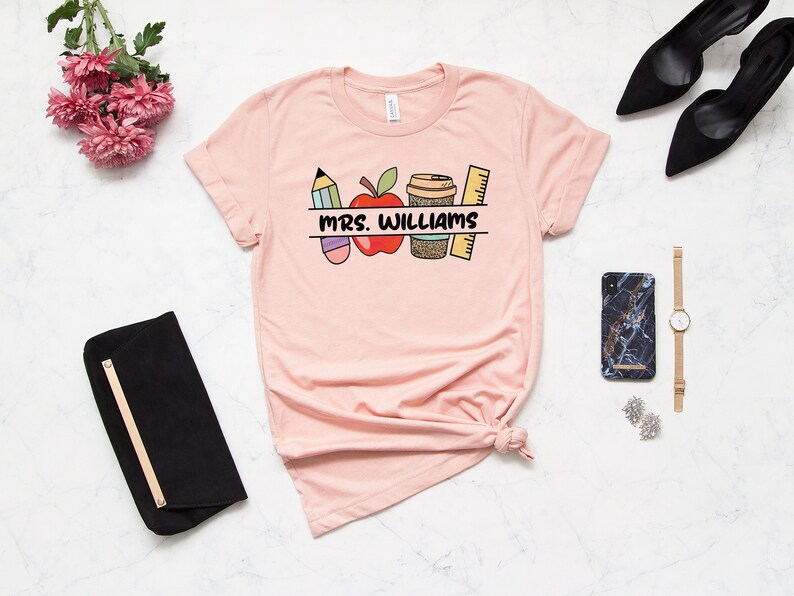 May include: A pink t-shirt with a graphic design of a pencil, apple, coffee cup, and ruler. The text "MRS. WILLIAMS" is printed on the shirt.