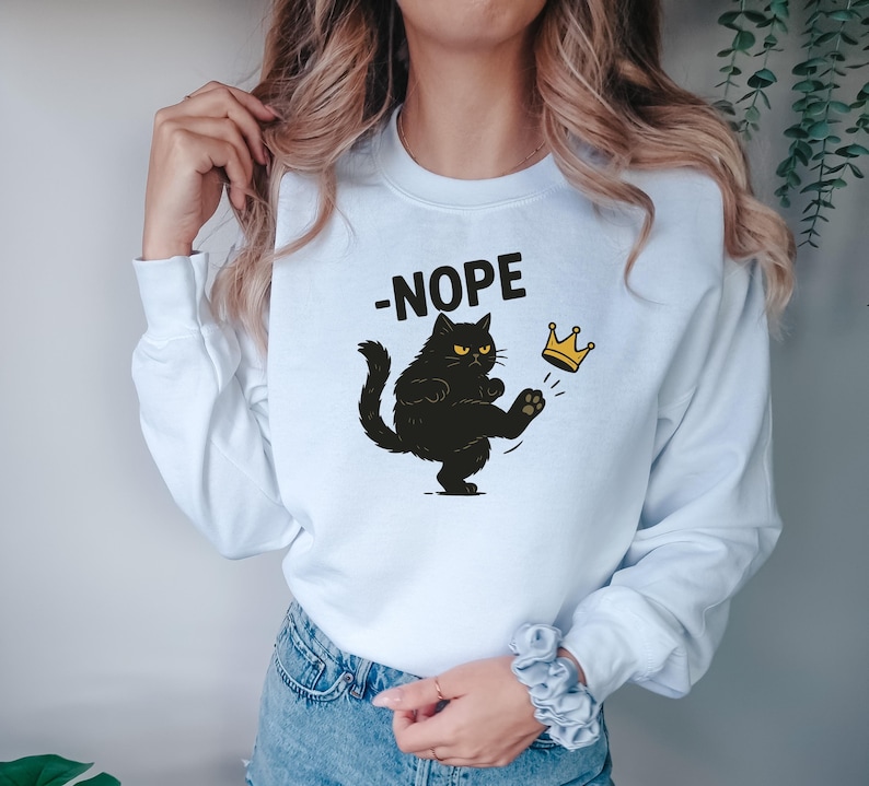 May include: White sweatshirt with a black cat graphic kicking a gold crown. The word "-NOPE" is printed above the cat. The cat has yellow eyes and is kicking the crown with its paw.