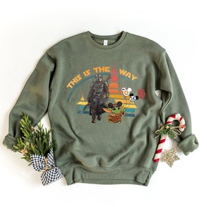 K&ouml;nnte beinhalten: Olivgr&uuml;nes Sweatshirt mit einer Grafik von The Mandalorian und Grogu, mit dem Text "THIS IS THE WAY". Das Design beinhaltet einen Retro-Sonnenuntergang und Mickey Mouse-Ballons. Ein festliches Urlaubsthema.