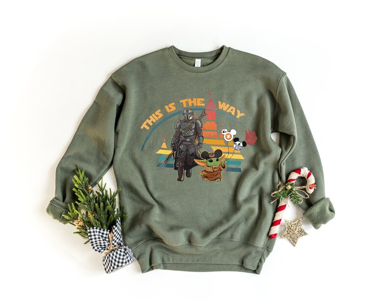 K&ouml;nnte beinhalten: Olivgr&uuml;nes Sweatshirt mit einer Grafik von The Mandalorian und Grogu, mit dem Text "THIS IS THE WAY". Das Design beinhaltet einen Retro-Sonnenuntergang und Mickey Mouse-Ballons. Ein festliches Urlaubsthema.