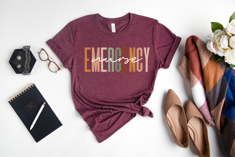 Puede incluir: Una camiseta de manga corta color granate con la palabra EMERGENCY en letras de bloque con colores tierra alternos y la palabra nurse escrita en cursiva blanca en el centro. La camiseta tiene cuello redondo y mangas remangadas.