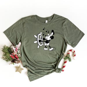May include: Olive green t-shirt featuring a black and white cartoon of a mouse operating a ship's wheel, with musical notes above. The shirt is styled with a knot at the bottom and is surrounded by holiday decorations.