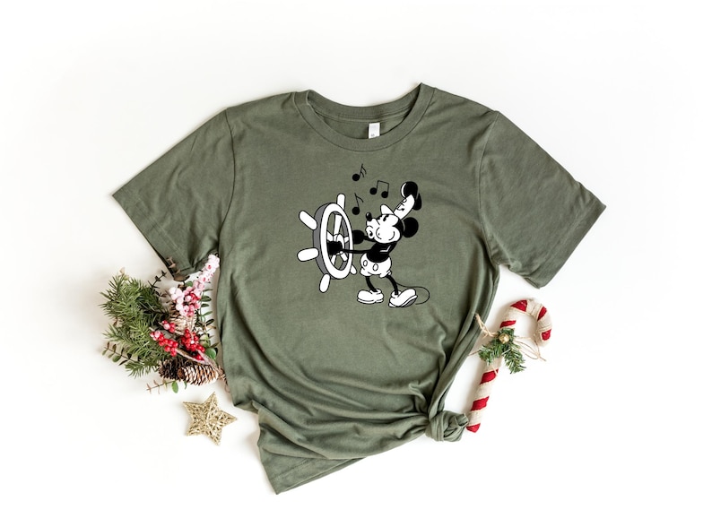 May include: Olive green t-shirt featuring a black and white cartoon of a mouse operating a ship's wheel, with musical notes above. The shirt is styled with a knot at the bottom and is surrounded by holiday decorations.