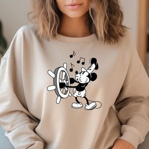 May include: Beige sweatshirt featuring a black and white graphic of Mickey Mouse at a ship's wheel, with musical notes. The design is centered on the front of the long-sleeved top.