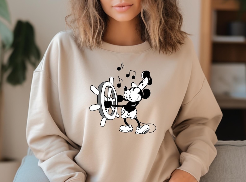 May include: Beige sweatshirt featuring a black and white graphic of Mickey Mouse at a ship's wheel, with musical notes. The design is centered on the front of the long-sleeved top.