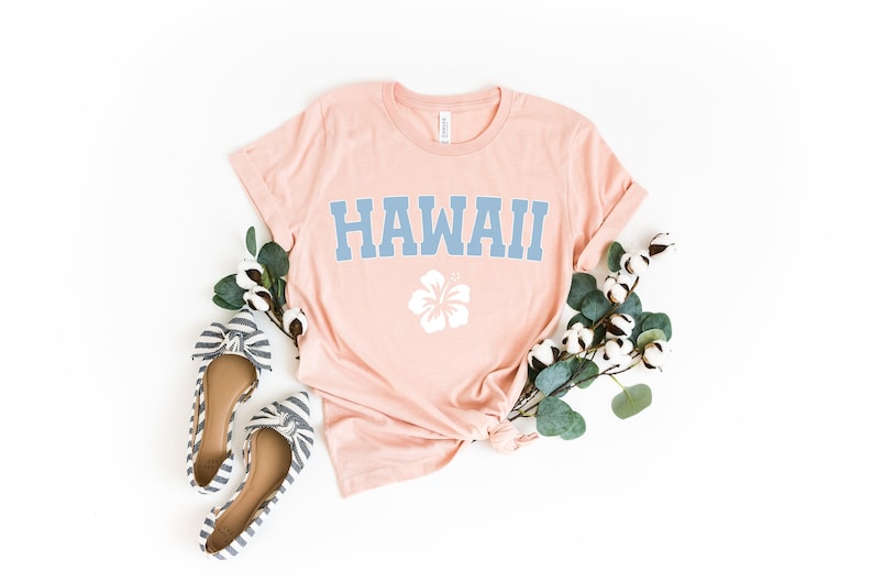 May include: A light pink t-shirt with the word "HAWAII" printed in blue and a white hibiscus flower graphic. The shirt is laid flat on a white background with a sprig of eucalyptus and cotton bolls.