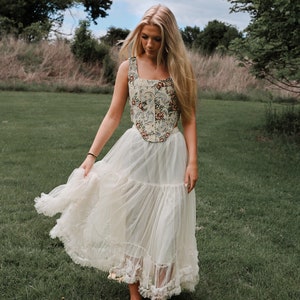 May include: A sleeveless corset top featuring a cream base with a multi-colored floral and scroll pattern, paired with a long, layered, off-white tulle skirt that has a ruffled hemline. The corset has a square neckline and a pointed front hem.
