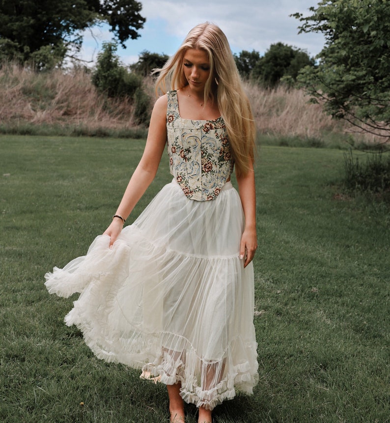 May include: A sleeveless corset top featuring a cream base with a multi-colored floral and scroll pattern, paired with a long, layered, off-white tulle skirt that has a ruffled hemline. The corset has a square neckline and a pointed front hem.