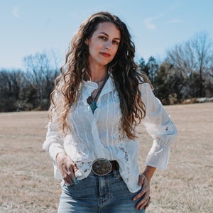 May include: A woman with long, curly brown hair wearing a white, lacy blouse with a turquoise necklace and a silver belt buckle. She is standing in a field with a blue sky in the background.