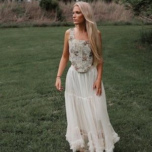 May include: A sleeveless corset top featuring a cream base with a multi-colored floral pattern in shades of red, blue, and green, paired with a floor-length, tiered white tulle skirt with ruffled hem detailing and a sheer, layered construction.