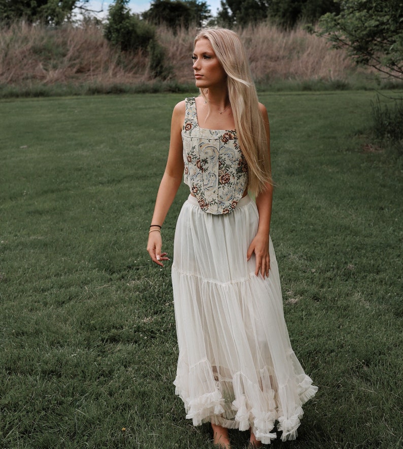 May include: A sleeveless corset top featuring a cream base with a multi-colored floral pattern in shades of red, blue, and green, paired with a floor-length, tiered white tulle skirt with ruffled hem detailing and a sheer, layered construction.