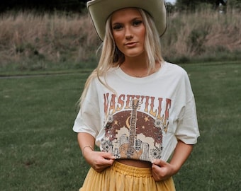 May include: A woman wearing a white t-shirt with a graphic of a guitar and the text "Nashville" on it. She is also wearing a yellow tulle skirt and a beige cowboy hat.
